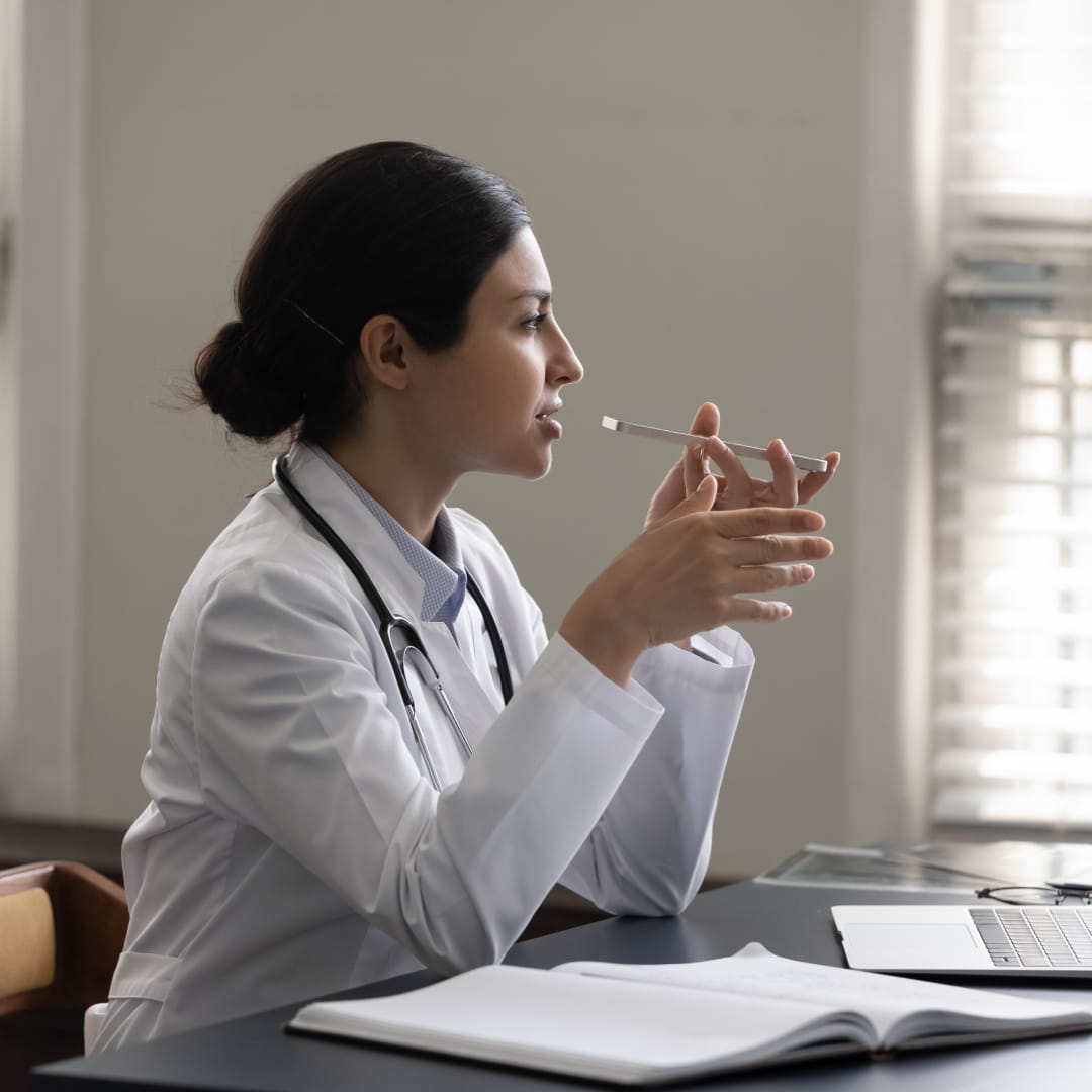 Doctor in white coat sits at desk, holding pen, looking contemplative, with open notebook and laptop in front of her.