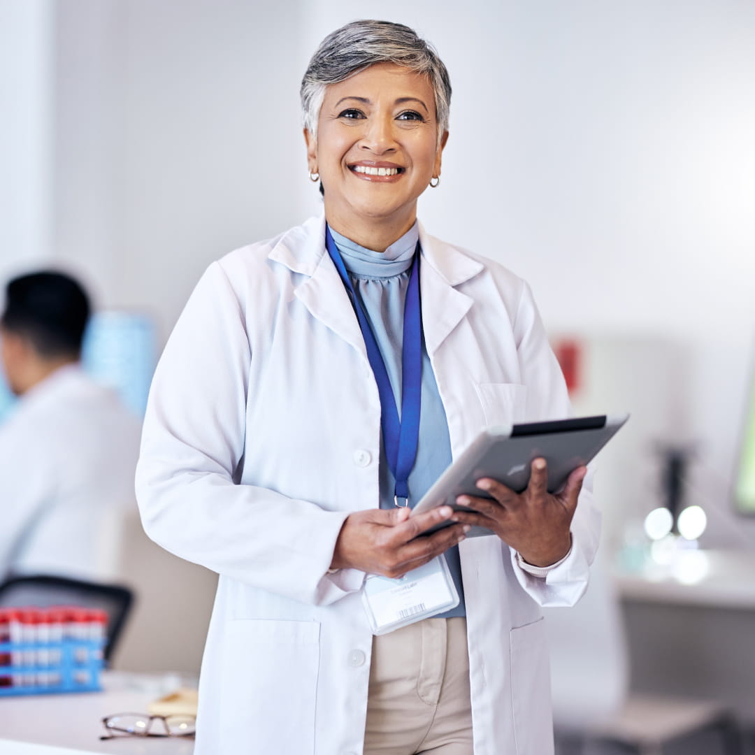 Senior female doctor in a white coat smiles while holding a tablet in a modern medical office setting.