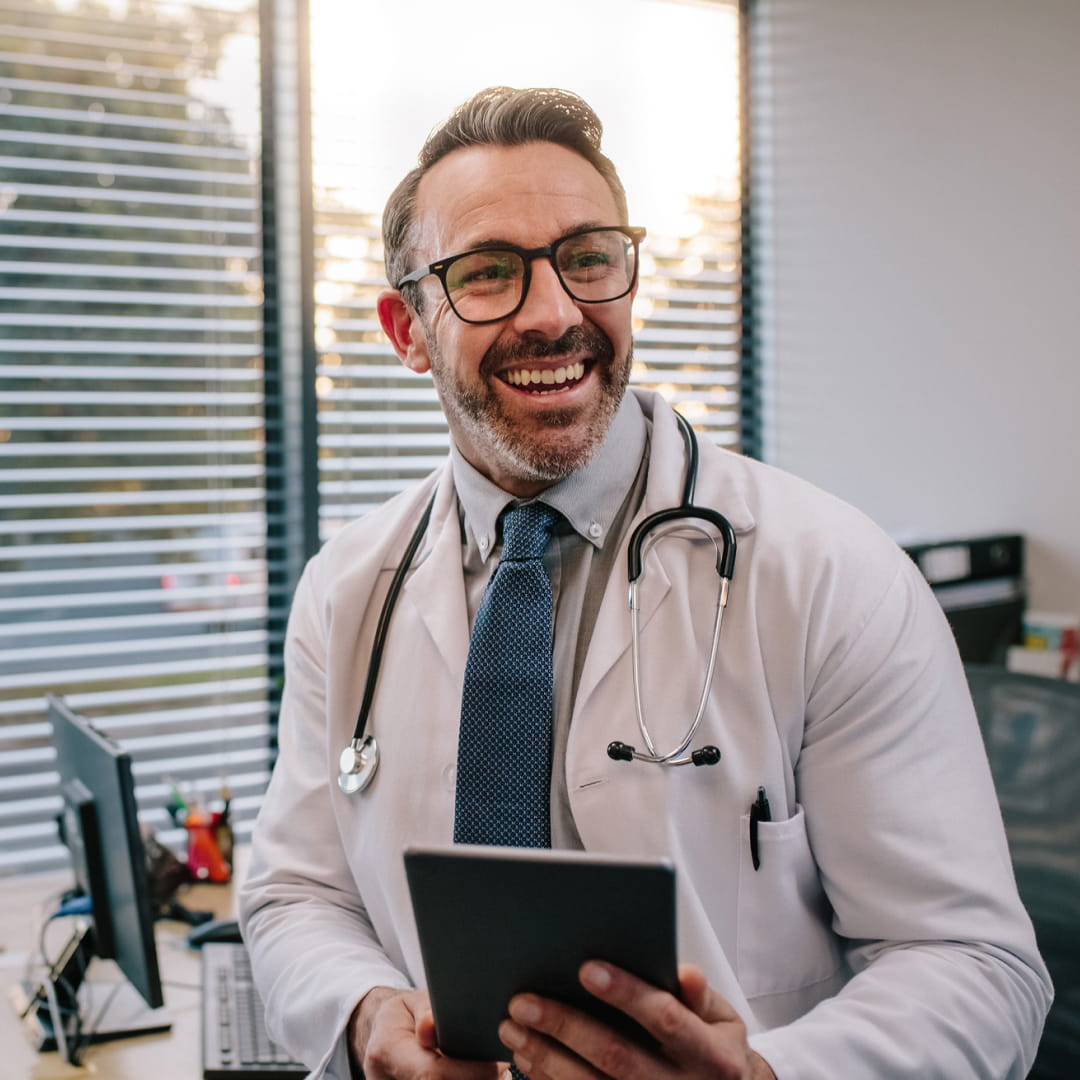 Smiling doctor with glasses and stethoscope holds a tablet in a sunlit office with blinds and computer desk.