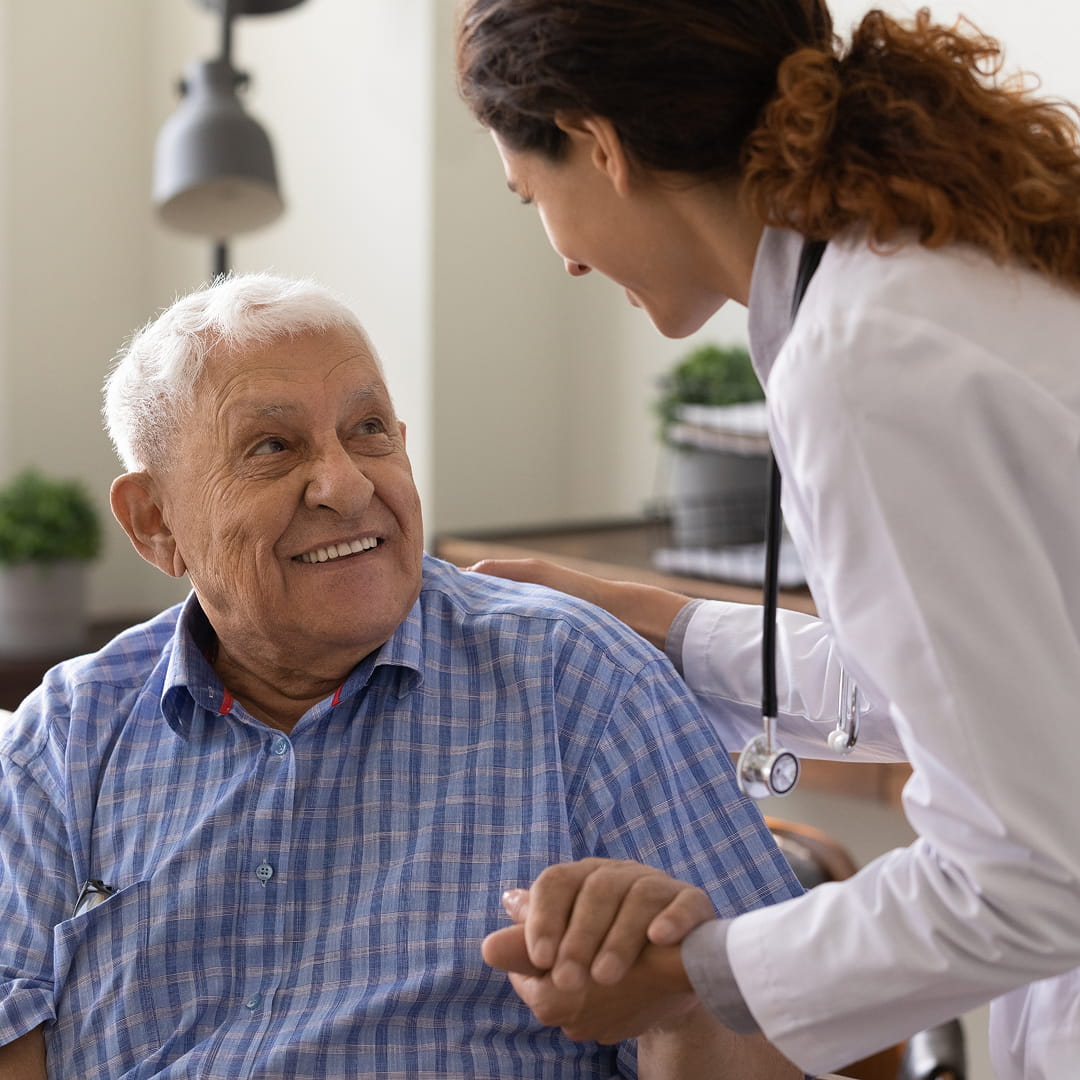 Elderly man smiling at female doctor holding his hand in a medical office setting.