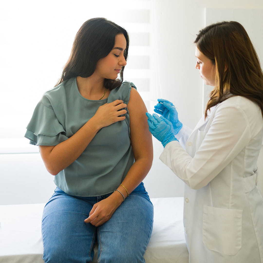 A woman receives a vaccine shot from a healthcare professional in a white coat and blue gloves in a bright room.