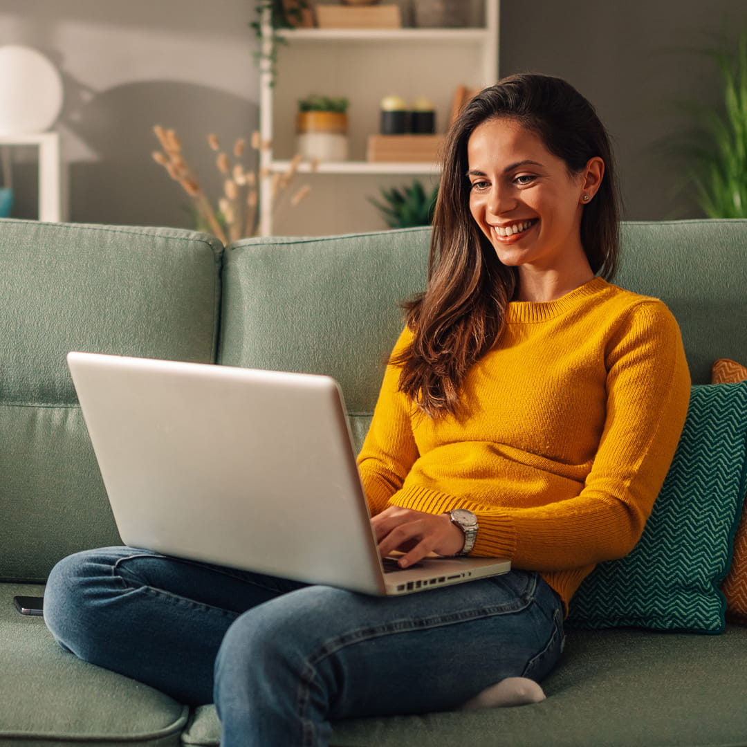 Woman in yellow sweater smiling while using a laptop on a green couch surrounded by plants and shelves.