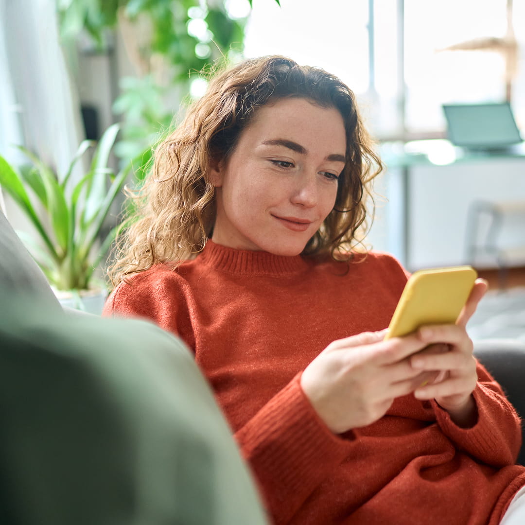 Person in an orange sweater uses a smartphone while sitting on a couch. Bright room with plants and a desk in the background.