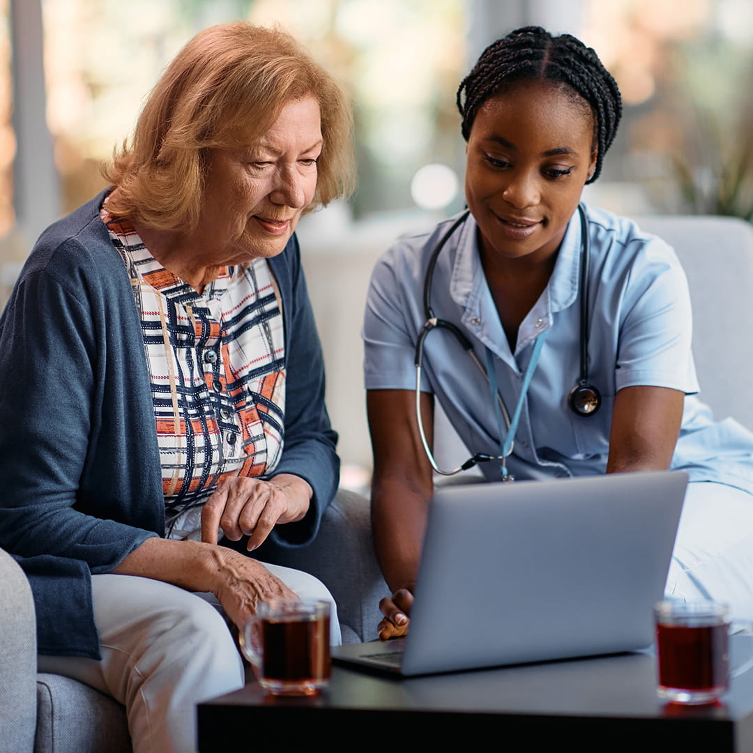 Elderly woman and nurse with a stethoscope look at a laptop together, seated in a well-lit room with cups of tea nearby.