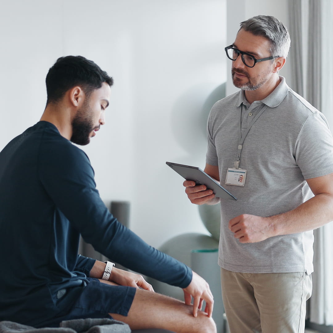 Physiotherapist with tablet consulting a patient sitting on a bench in a well-lit room.