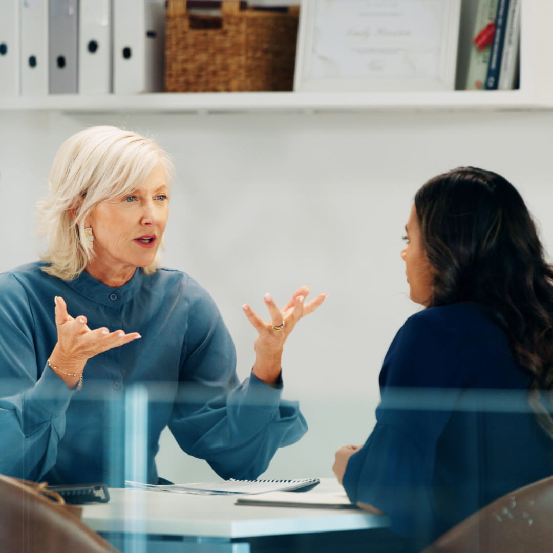 Two women engaged in a discussion at an office table; one gestures expressively with open hands.