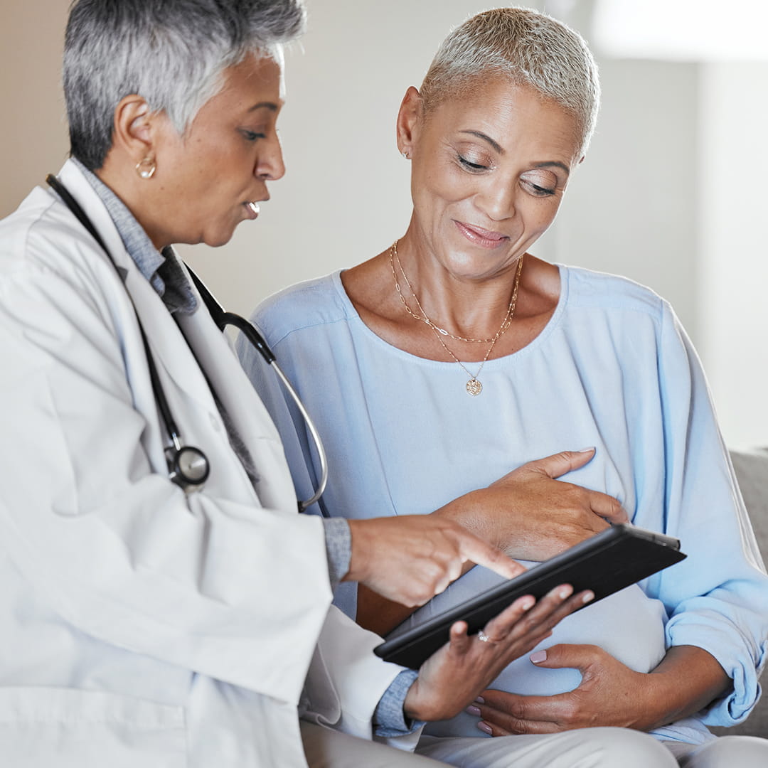 Doctor showing a tablet to a seated woman, both with short gray hair, in a clinical setting.