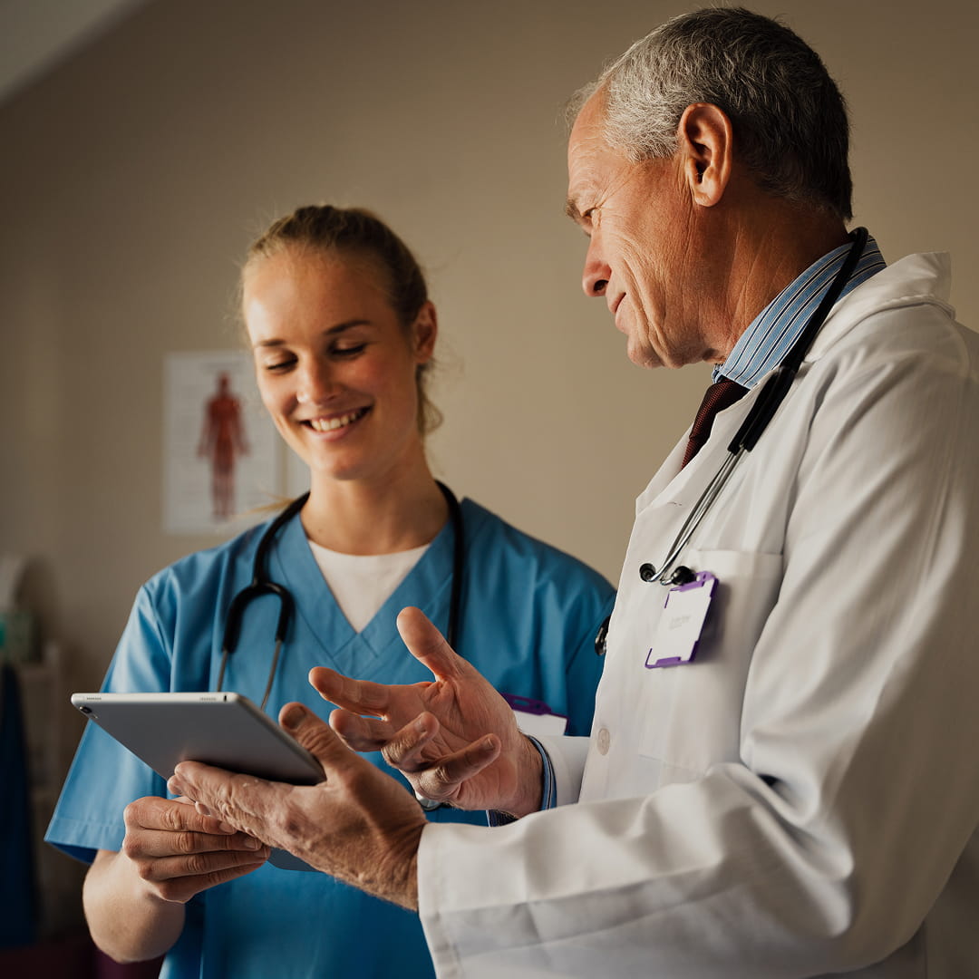 Doctor and nurse discussing while looking at a tablet in a clinic, both smiling, with a poster of a human figure in the background.