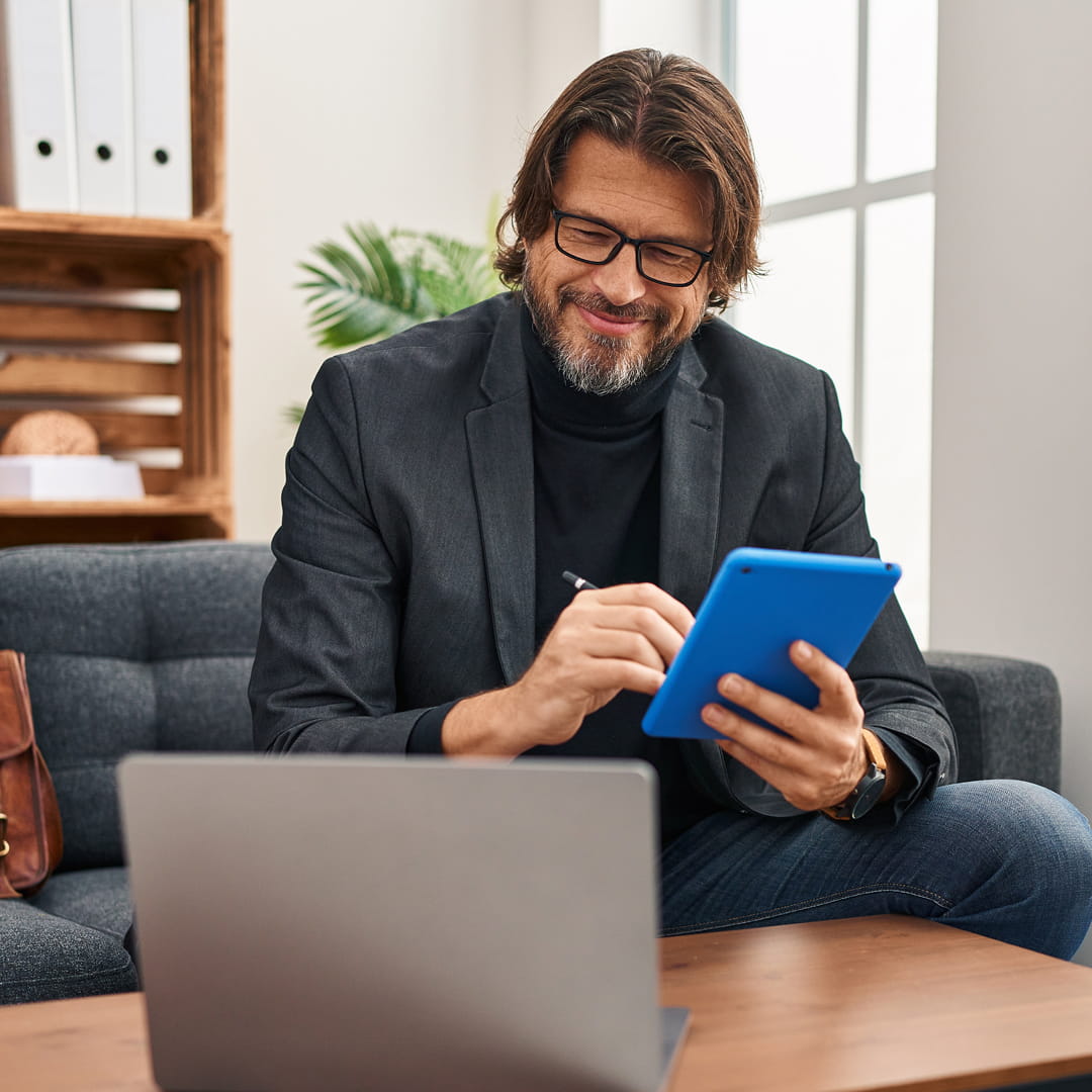 Man with glasses and long hair smiles while using a blue tablet pen. Seated on a sofa, laptop open, bookshelves in the background.