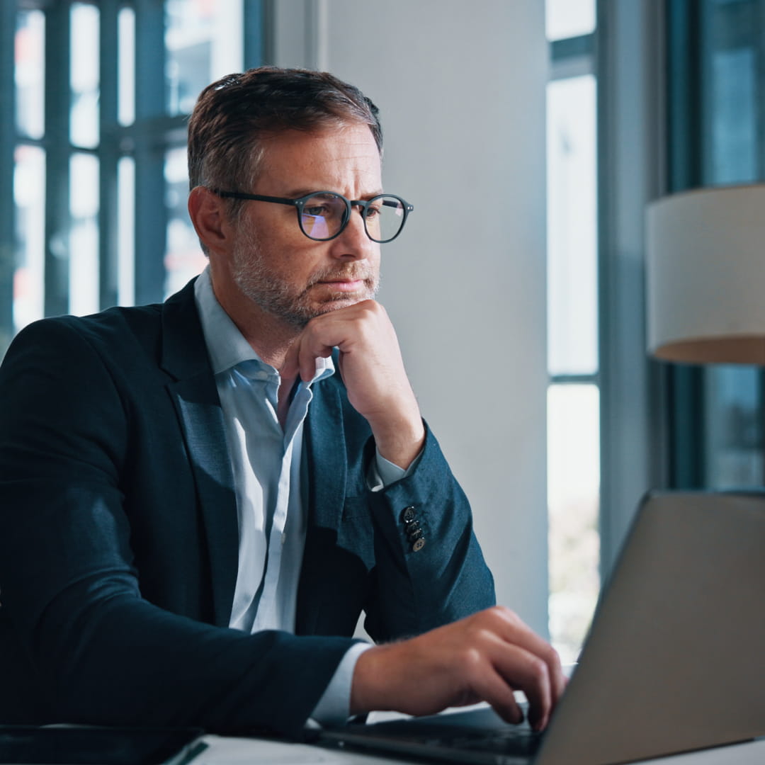Man in a suit wearing glasses works on a laptop in a modern office, resting his chin on his hand thoughtfully.