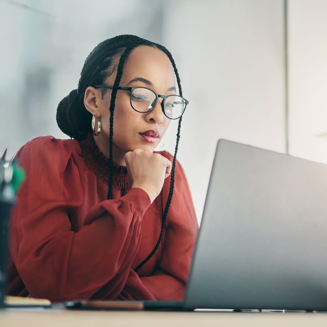 Person with braided hair and glasses wearing a red blouse, focused on a laptop in an office setting.