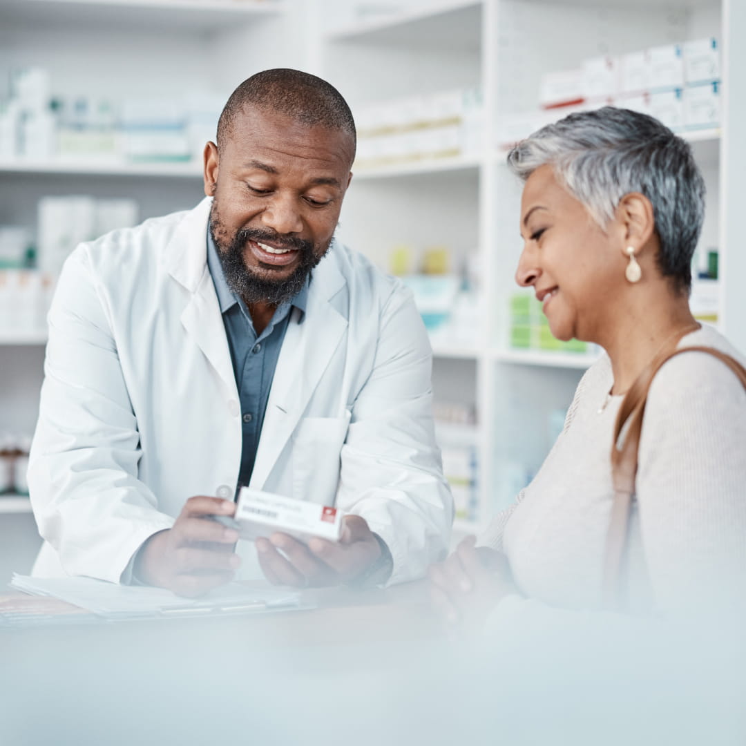 Pharmacist in a white coat explaining medication to a smiling woman in a pharmacy with shelves of products in the background.