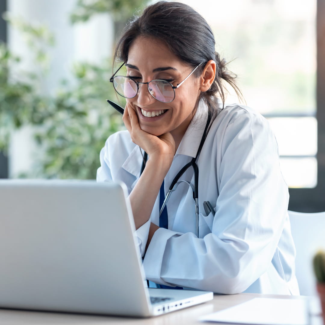Doctor smiling while looking at a laptop, sitting at a desk with plants in the background, wearing glasses and a stethoscope.