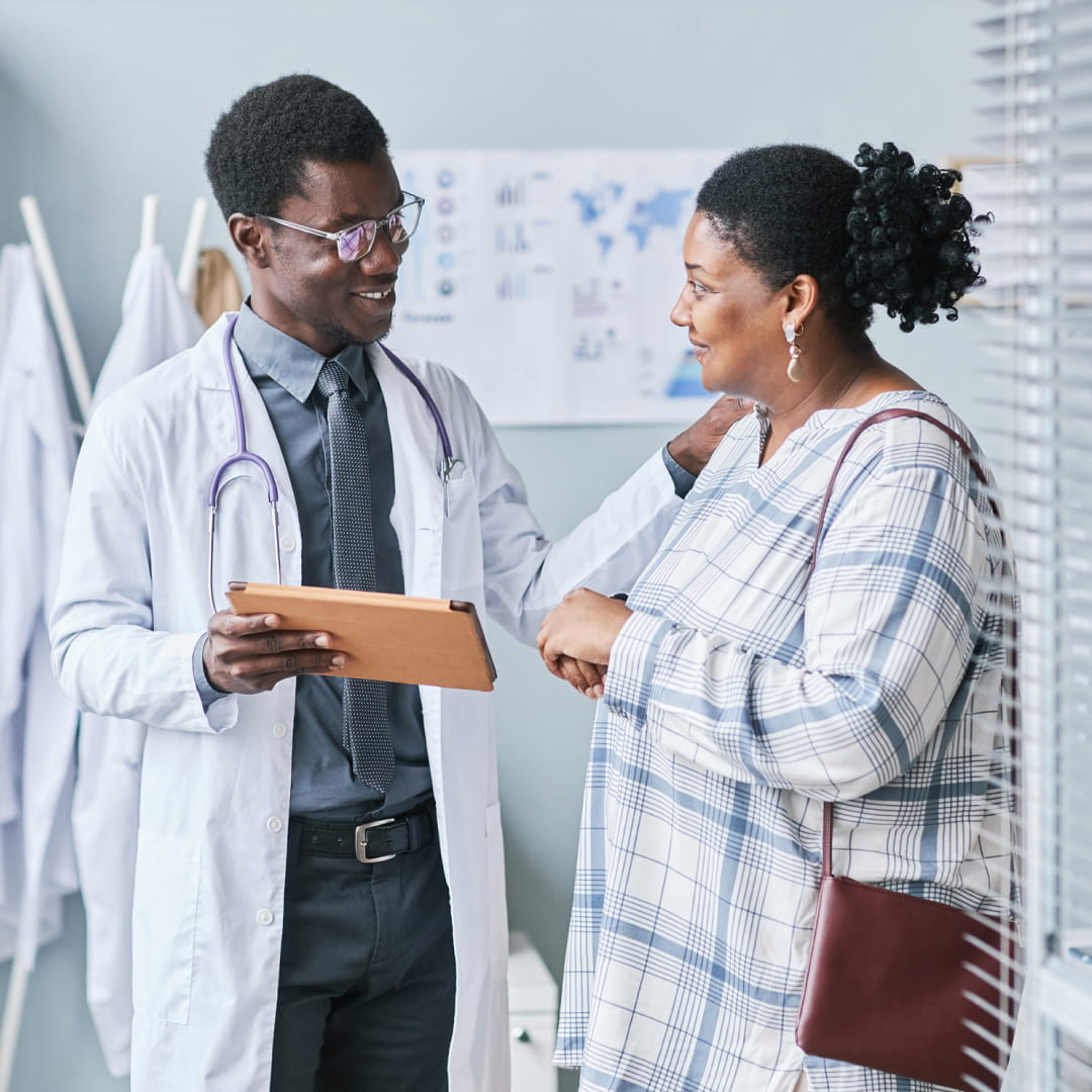 Doctor with clipboard smiles and talks to woman in patterned dress, touching her shoulder in a medical office.