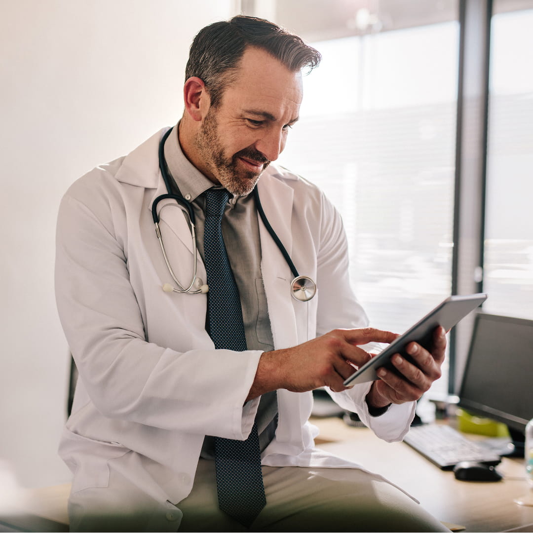 Doctor in white coat with stethoscope uses tablet in bright office, smiling and concentrating on screen.