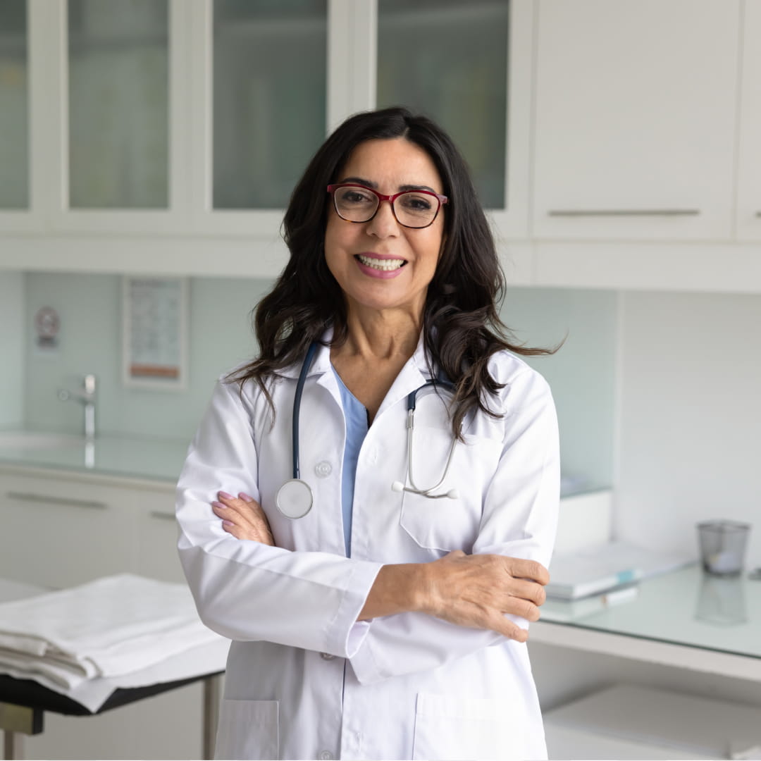 A smiling woman in a white coat and stethoscope stands in a bright medical office with arms crossed.