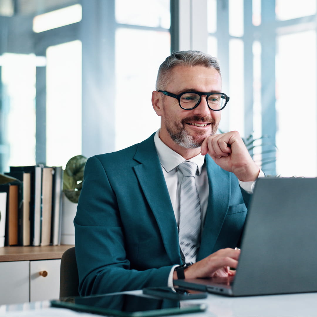 Man with glasses and beard working on a laptop in an office, wearing a suit, hand on chin, looking at screen thoughtfully.