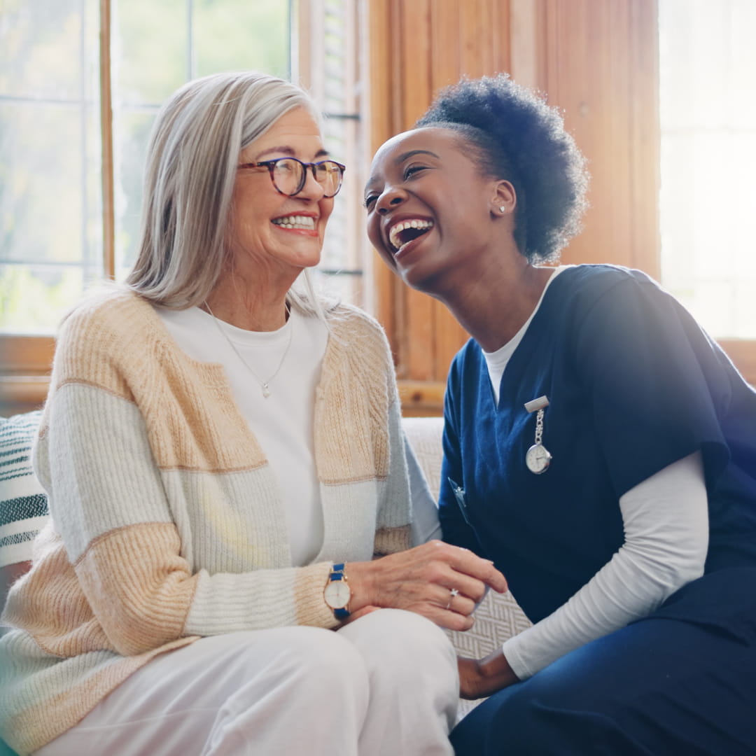 Elderly woman and caregiver in blue scrubs sharing a joyful moment, sitting on a couch by a window.
