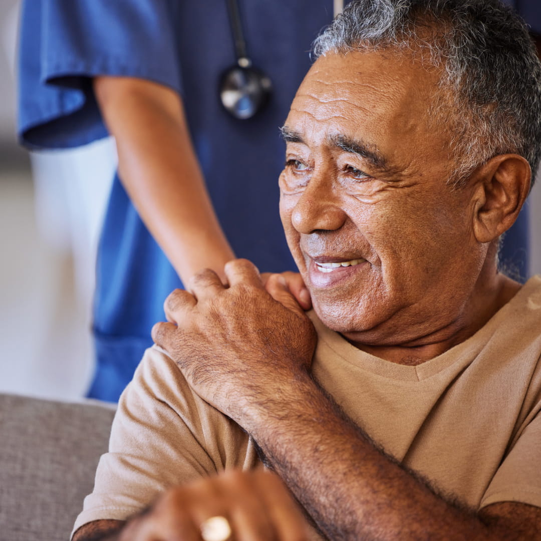 Elderly man smiling with a nurse in blue scrubs gently touching his shoulder in a supportive gesture.