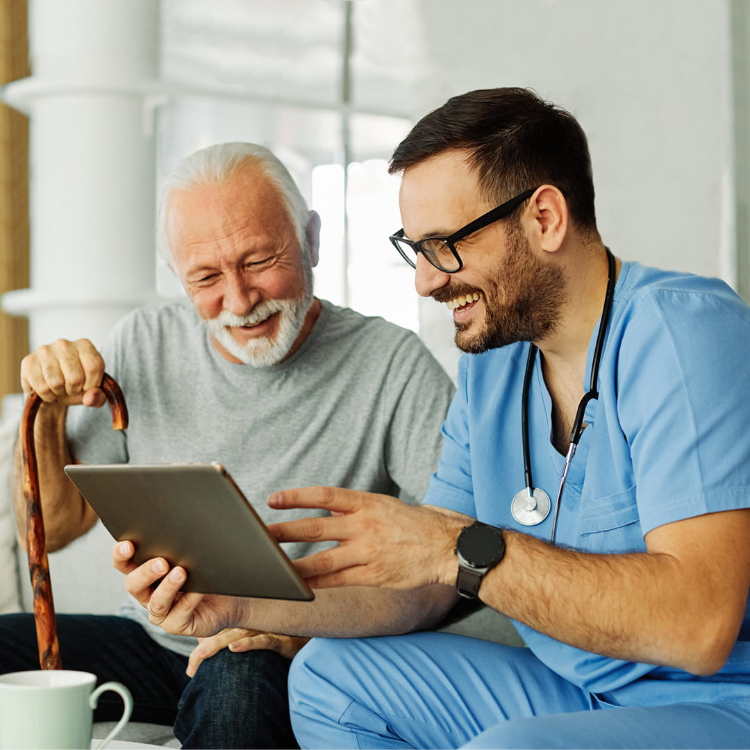 Elderly man with cane and young man in scrubs smiling while looking at a tablet in a bright room.
