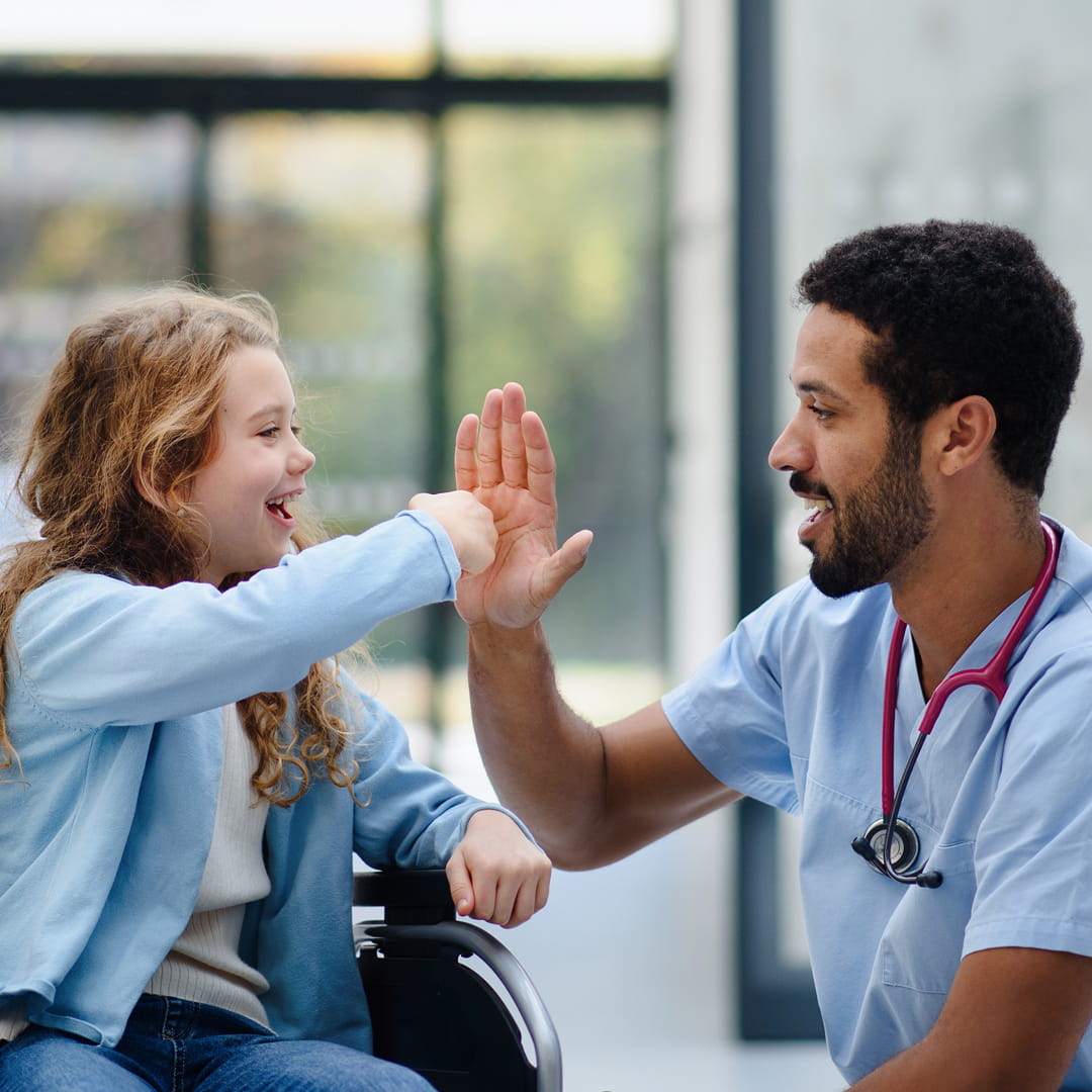 Girl in wheelchair high-fives smiling nurse in scrubs with stethoscope, in a bright, modern room.