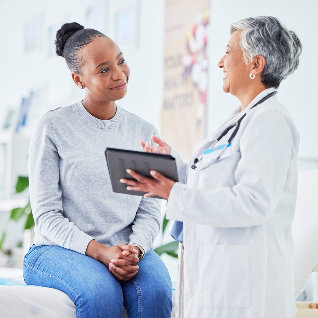 Doctor holding a tablet talks to a smiling patient sitting on an examination table in a medical office.