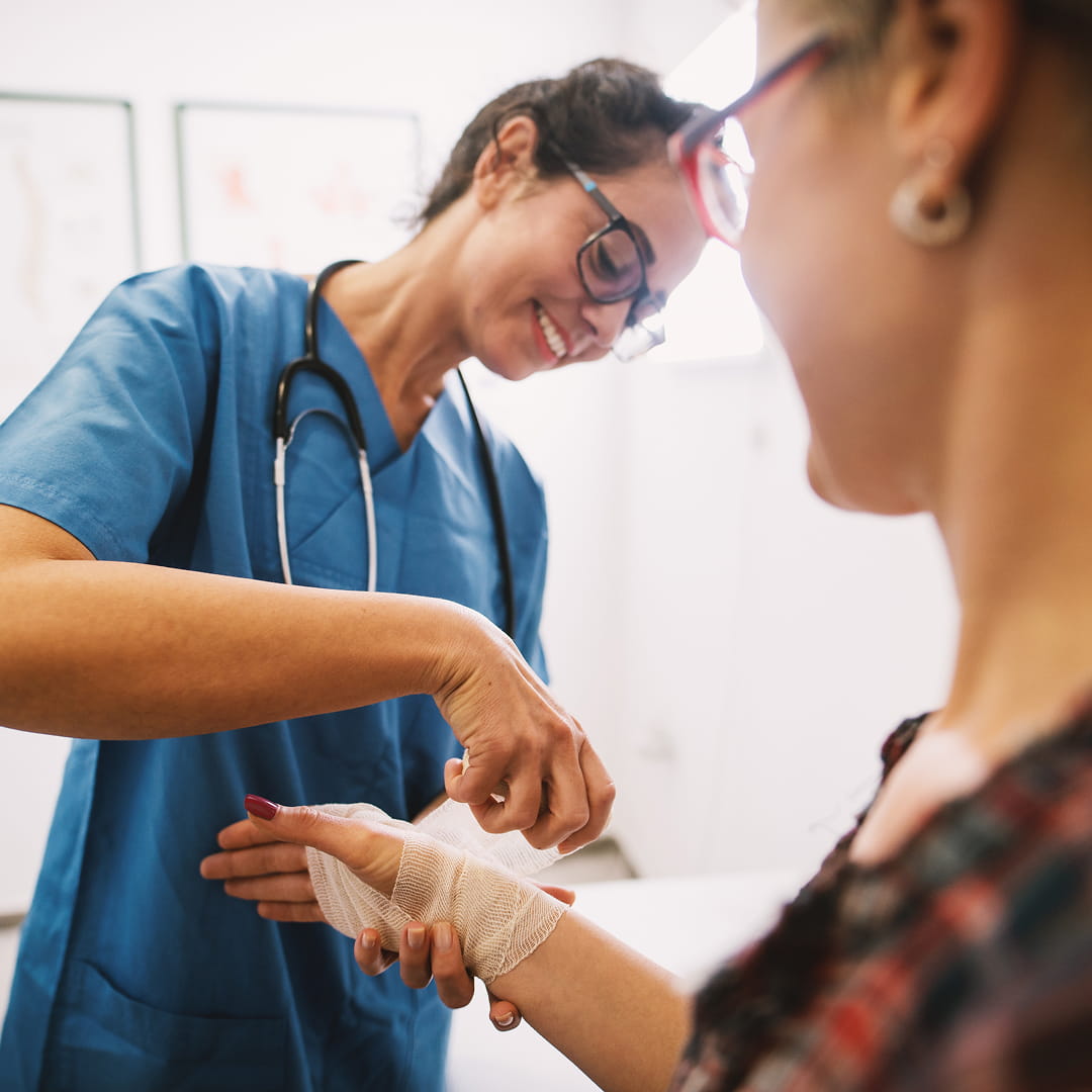 Nurse in blue scrubs bandaging a patient's hand in a medical office with charts on the wall.