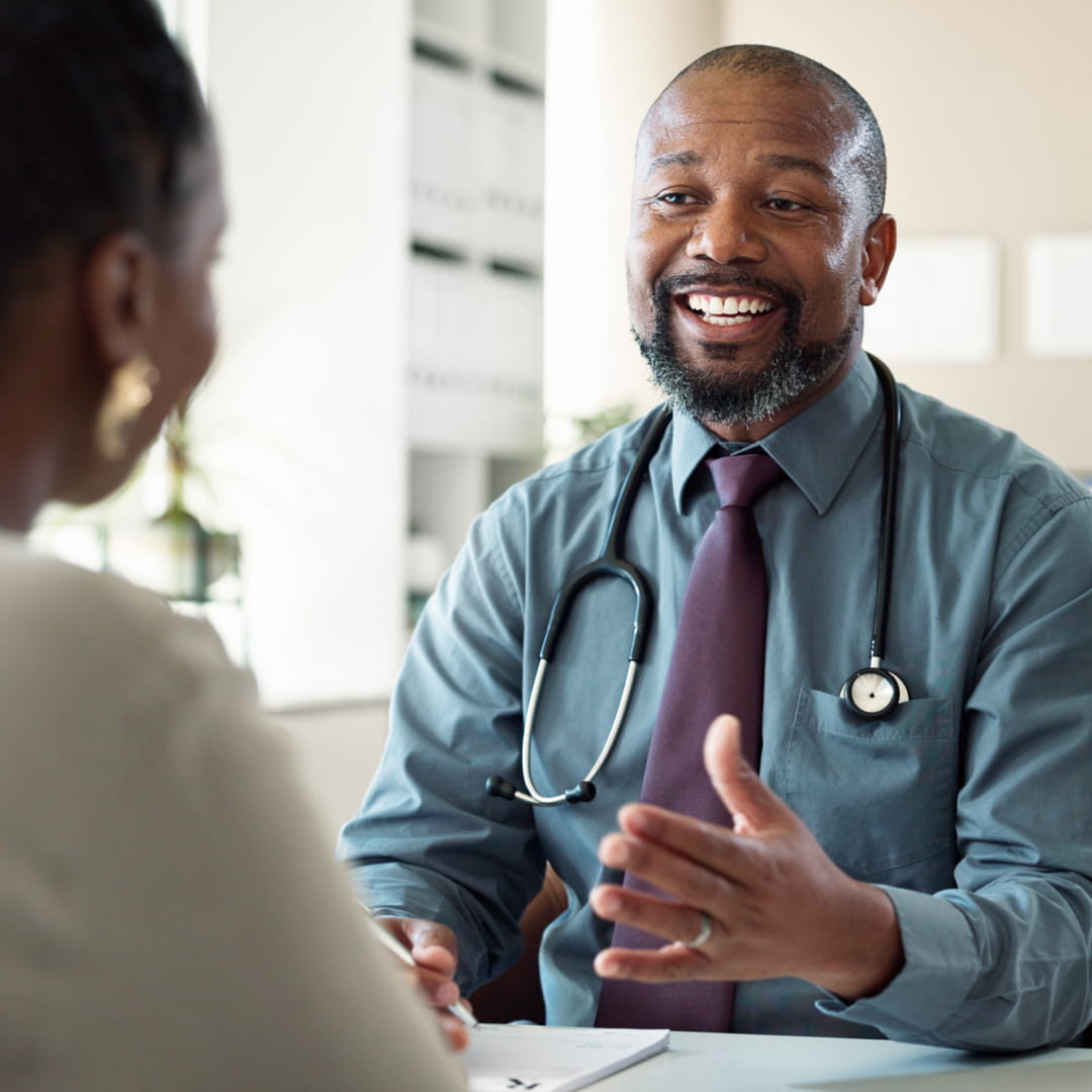 Doctor with stethoscope, smiling and speaking to a patient in an office setting, while gesturing with a hand.
