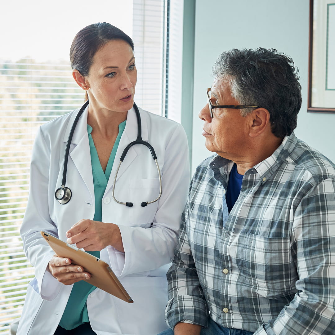 Doctor with stethoscope discussing with an older man in a plaid shirt, holding a tablet in a well-lit office.