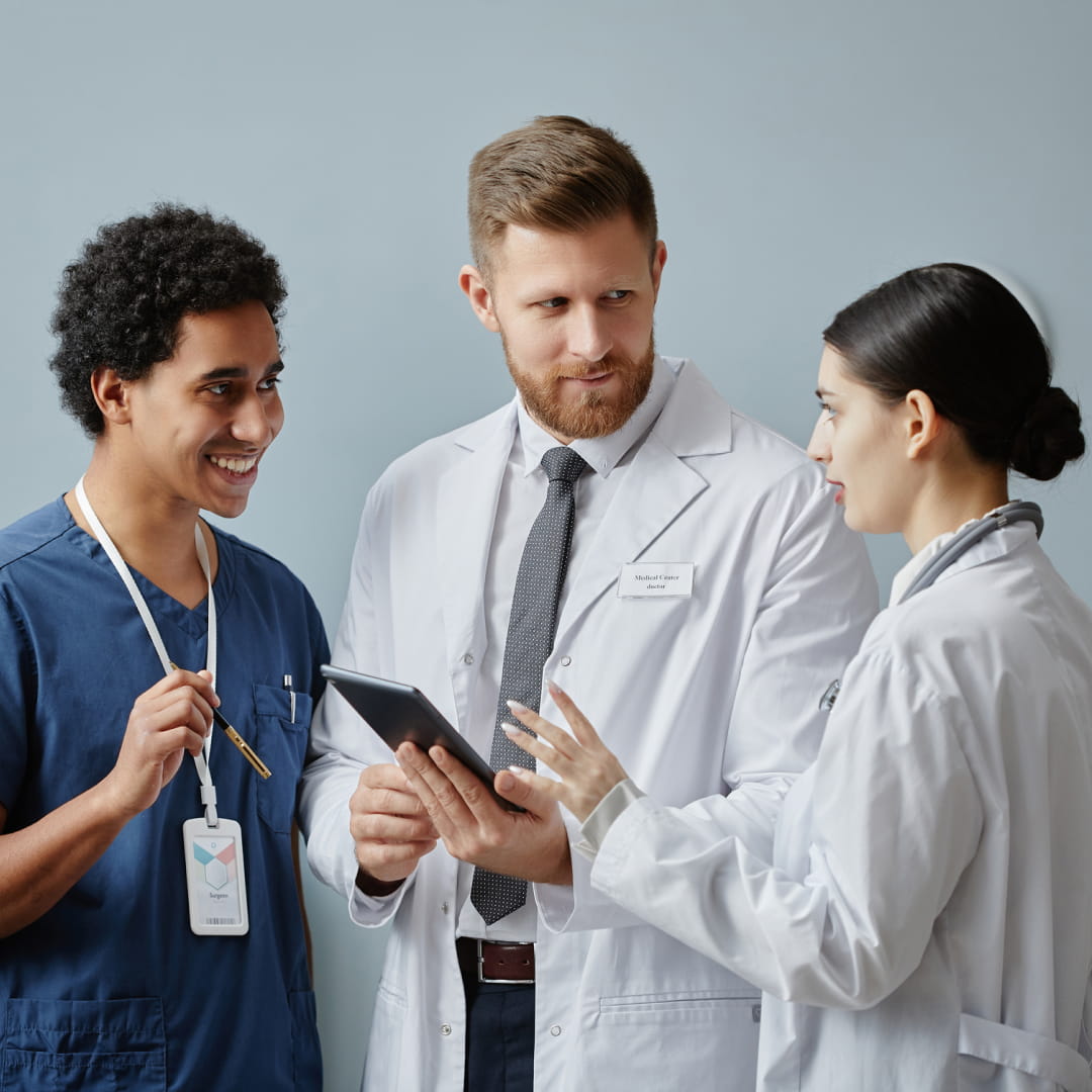 Medical professionals in discussion: a man in a lab coat with tablet, a woman in a lab coat gesturing, man in scrubs smiling.