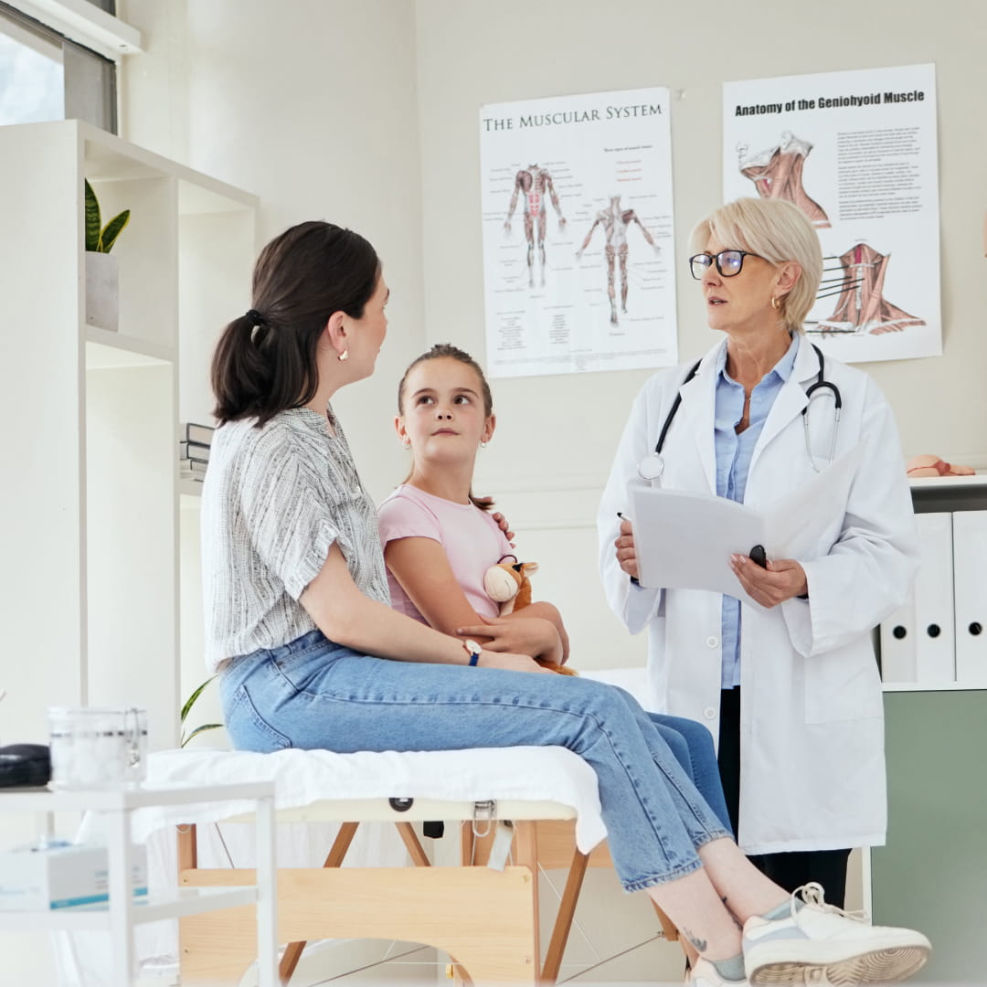Doctor in a white coat talks to a woman and child sitting on an exam table in a medical office with anatomy posters.