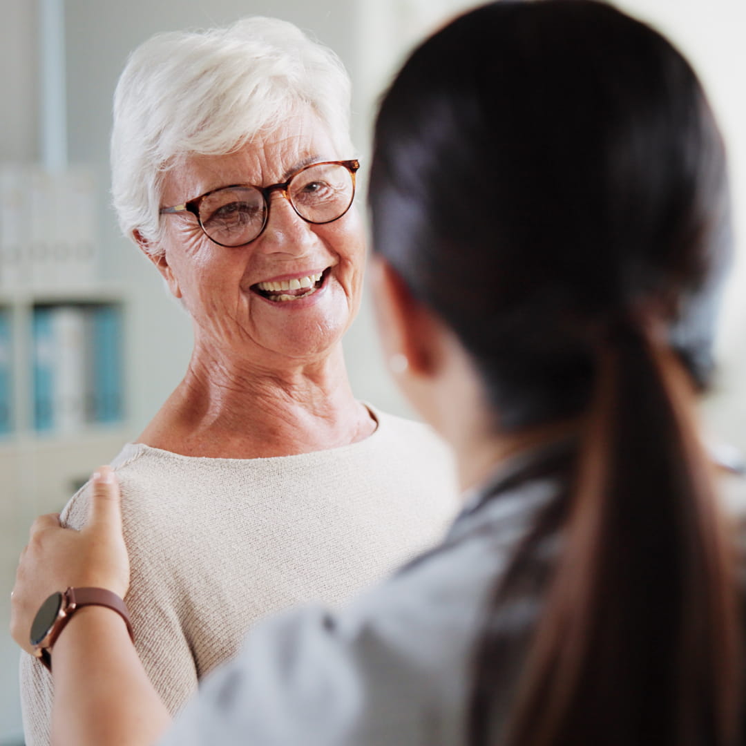 Elderly woman with white hair and glasses smiling at a person with a ponytail, who is touching her shoulders gently.