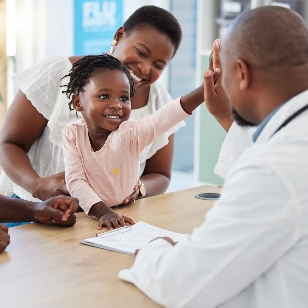 Child high-fiving a doctor at a clinic while sitting on a woman's lap, with a flu shot poster in the background.