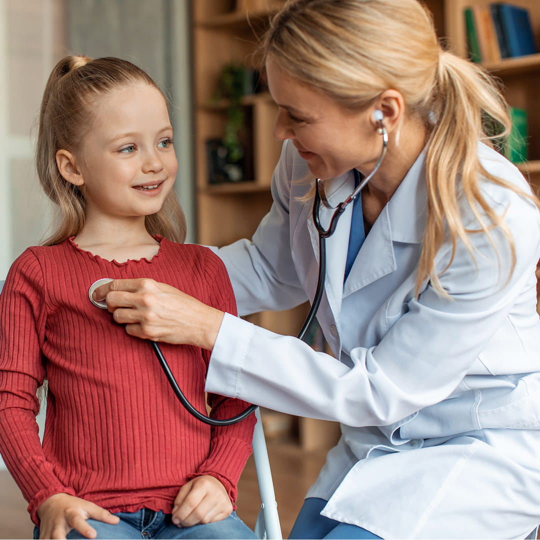 Doctor with stethoscope examines smiling girl in a red shirt, seated in a medical office with shelves in the background.