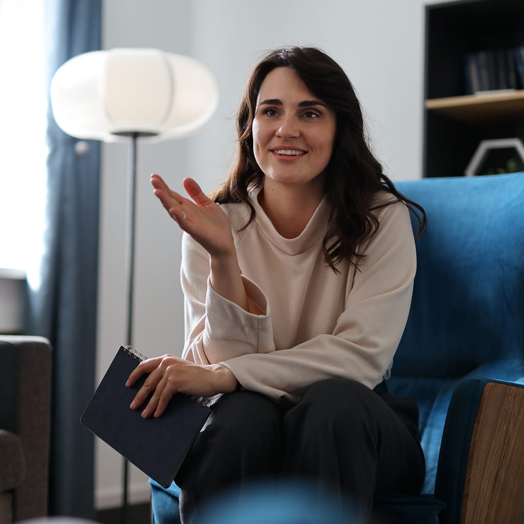 A woman with dark hair gestures while holding a clipboard, sitting on a blue chair in a modern, well-lit room with a lamp.