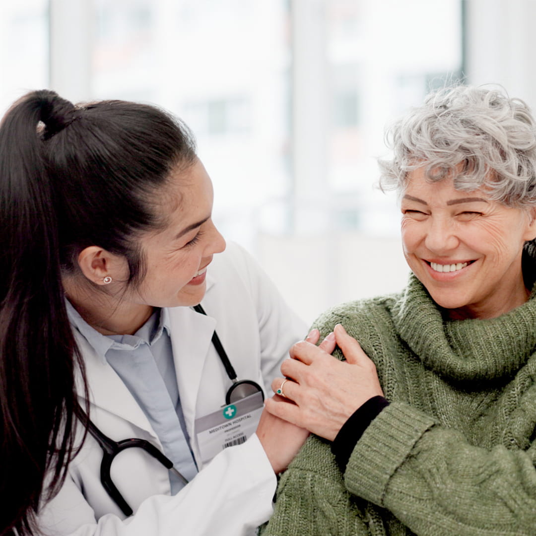 Doctor smiling with a stethoscope speaks with a laughing woman in a green sweater in a bright room.