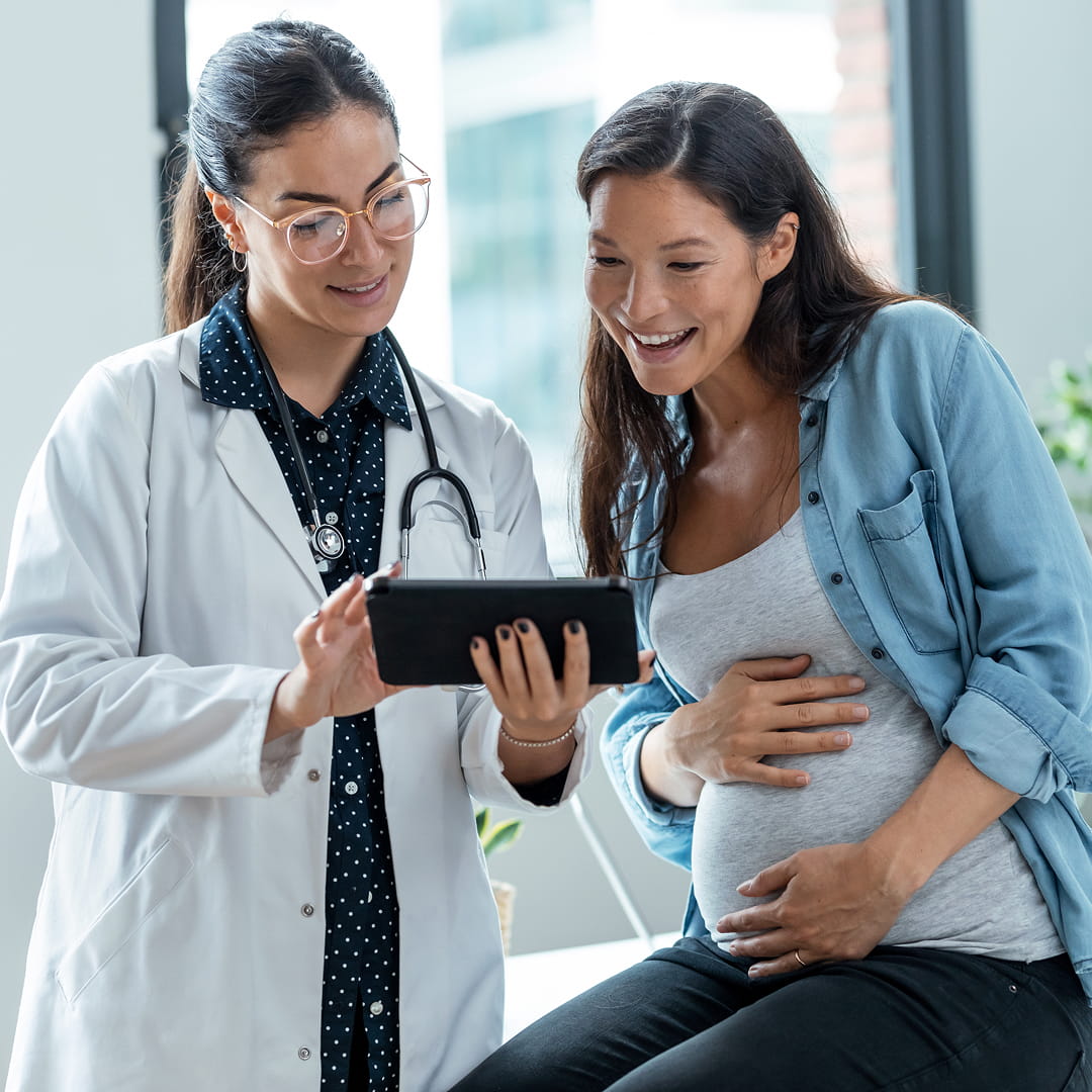 Doctor showing a pregnant woman something on a tablet. The woman is smiling and touching her belly.