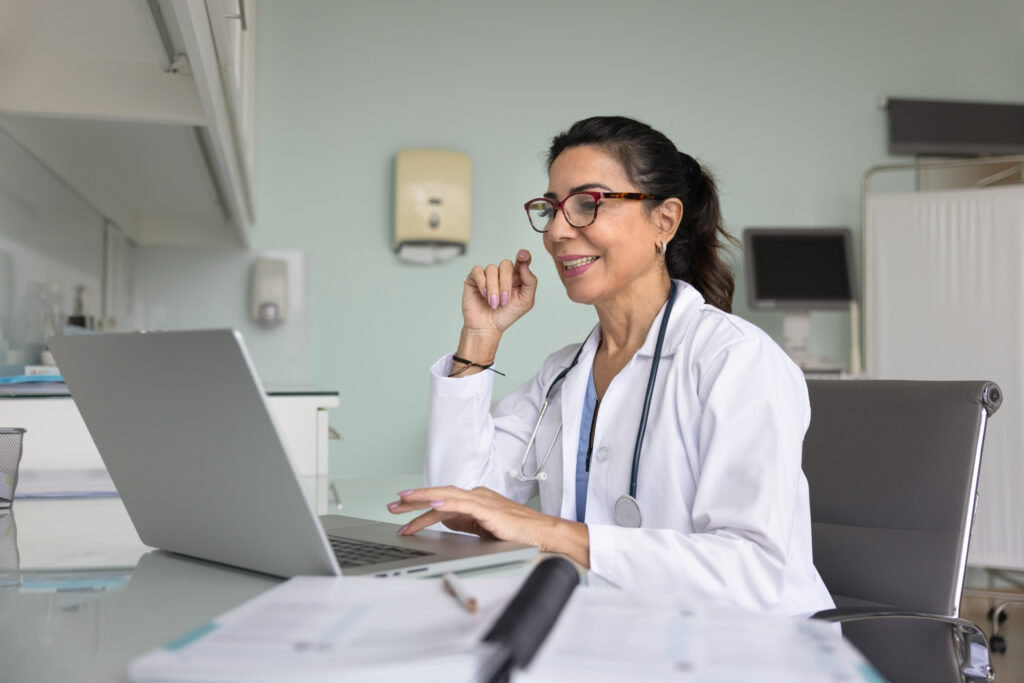 Positive happy mature physician doctor woman working at laptop in hospital office, typing, smiling, talking on video telemedicine conference call to patient, using Internet communication