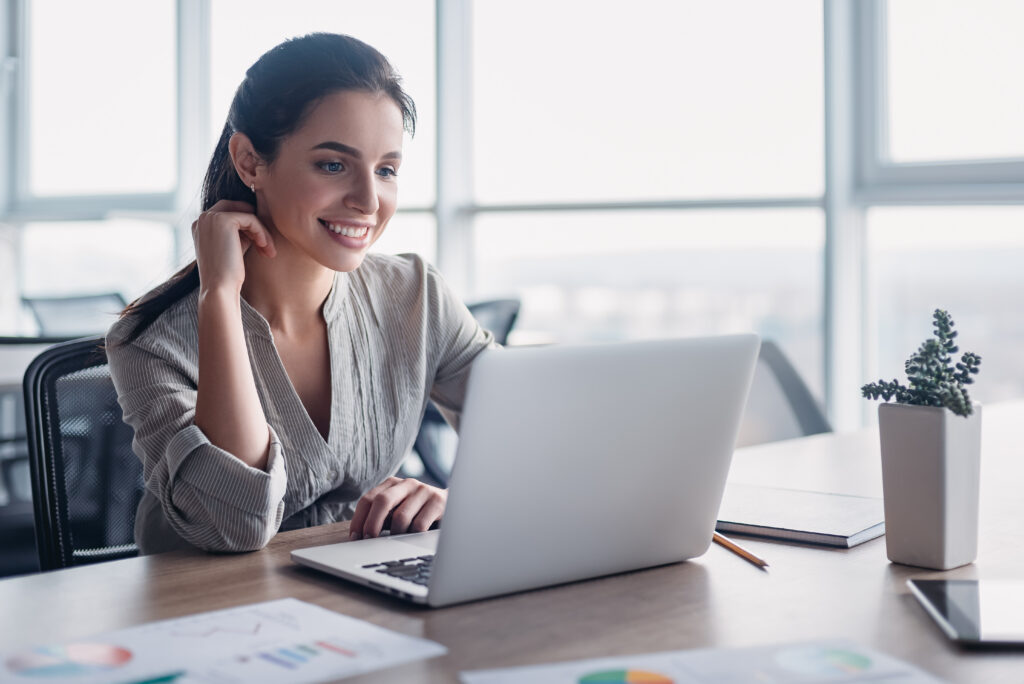 Young dark-haired businesswoman wearing striped shirt is sitting in her office and looking into laptop. Her phone, notebook and pencil are lying on the desk. Bright modern office.