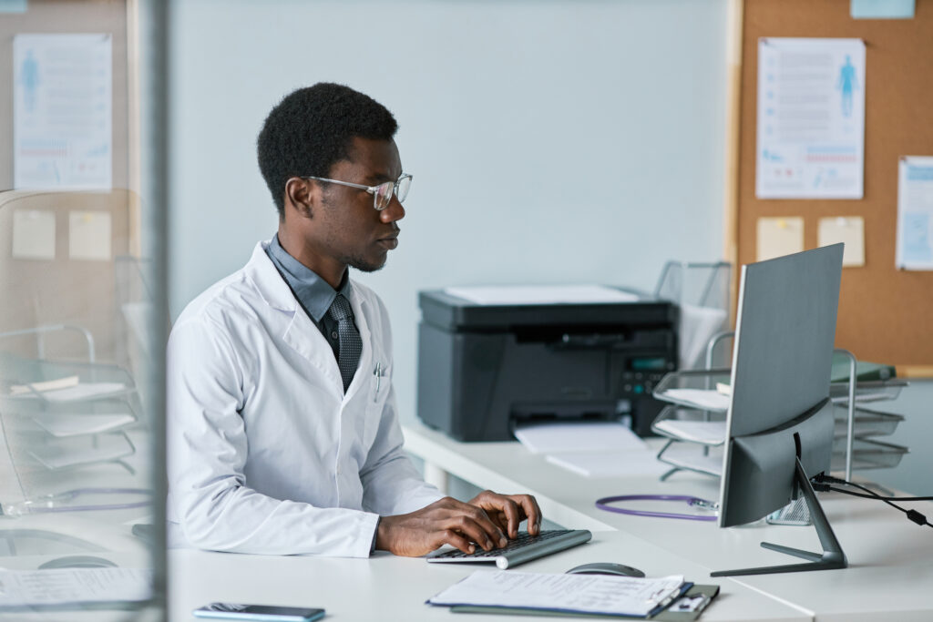 Side view portrait of young black doctor using computer at desk in office while working in private clinic