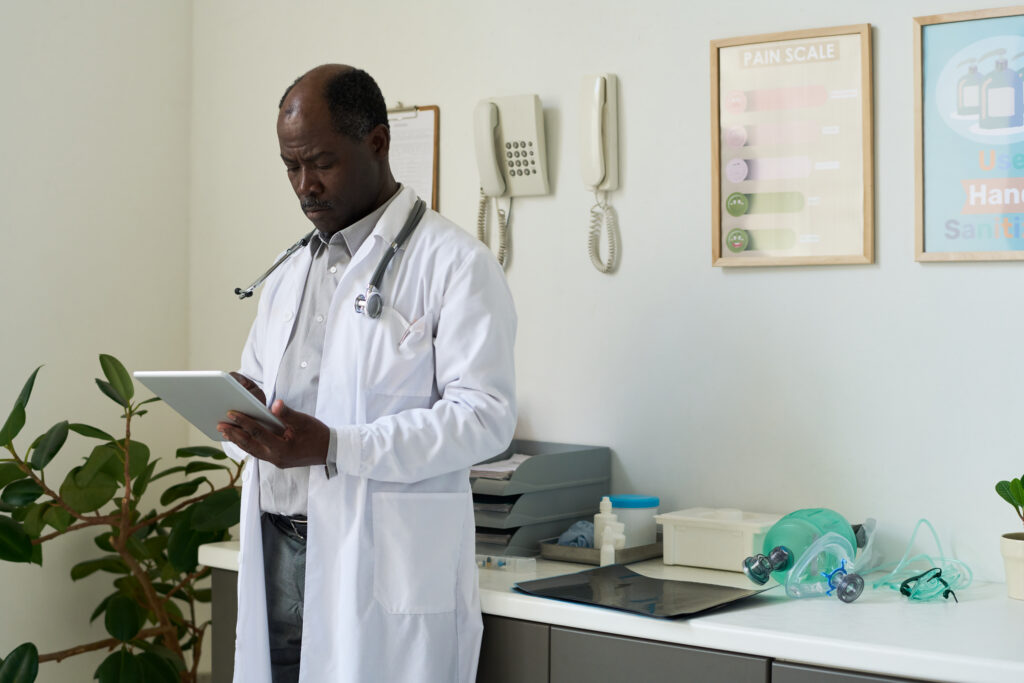 African American male doctor wearing lab coat and stethoscope, using tablet device while standing in medical office surrounded by medical equipment and plants