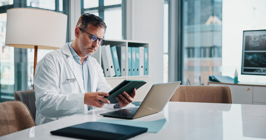 Healthcare, laptop and tablet with doctor man at desk in office of hospital for diagnosis, planning or research. Computer, medical and technology with mature medicine professional reading in clinic.