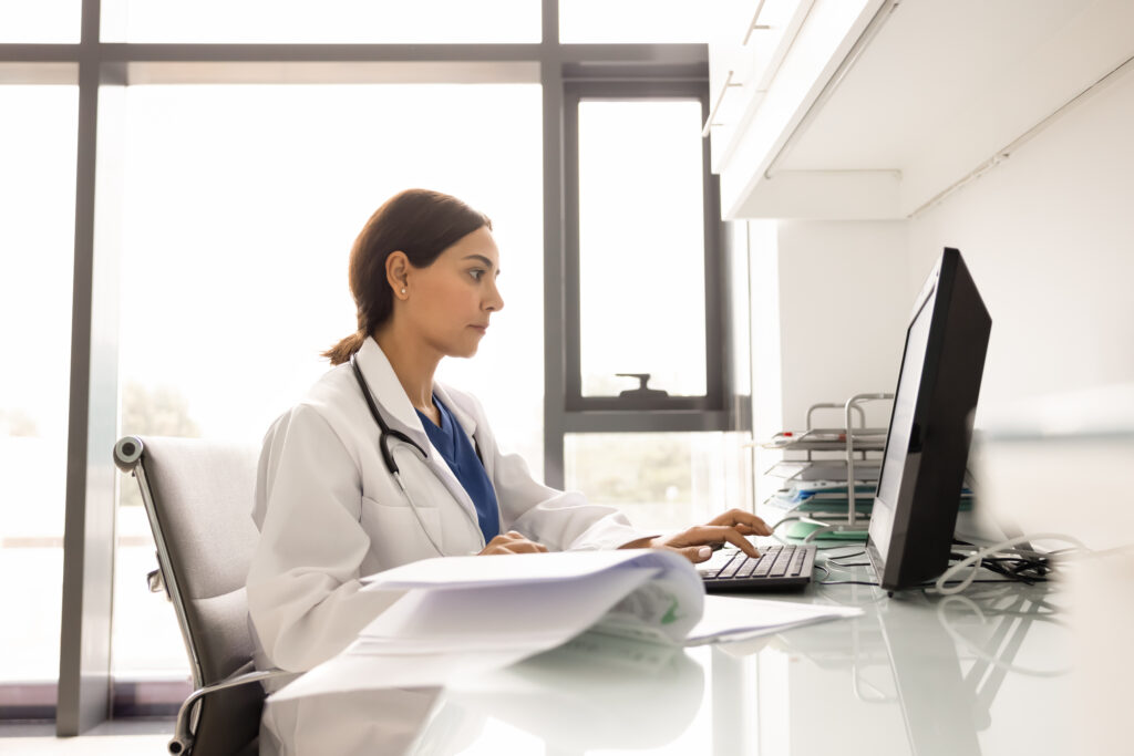 Focused female doctor in white coat working on desktop computer at modern clinic desk, surrounded by documents, history journals and anamnesis. Patient record management, medical research, healthcare