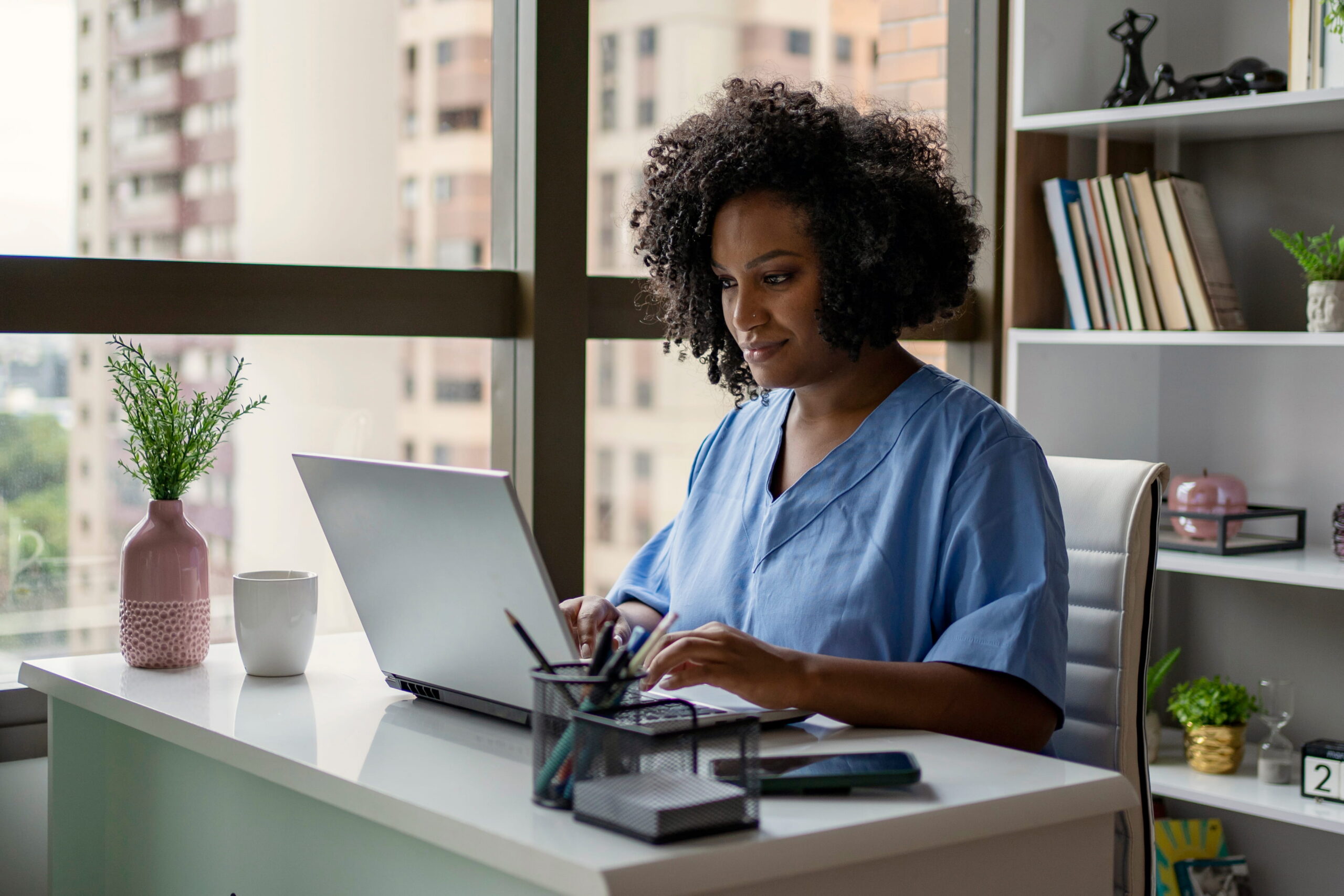 Healthcare professional in blue scrubs working on a laptop at her office desk